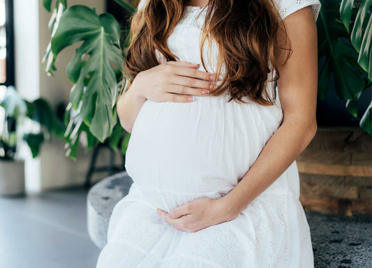 A zoomed-in picture of a woman holding her pregnant belly. She is in a white dress and sitting on a round bench. Pregnancy