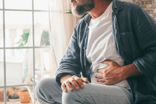 Man sits by the window one hand on his knee and the other holding his stomach in pain. Prostate